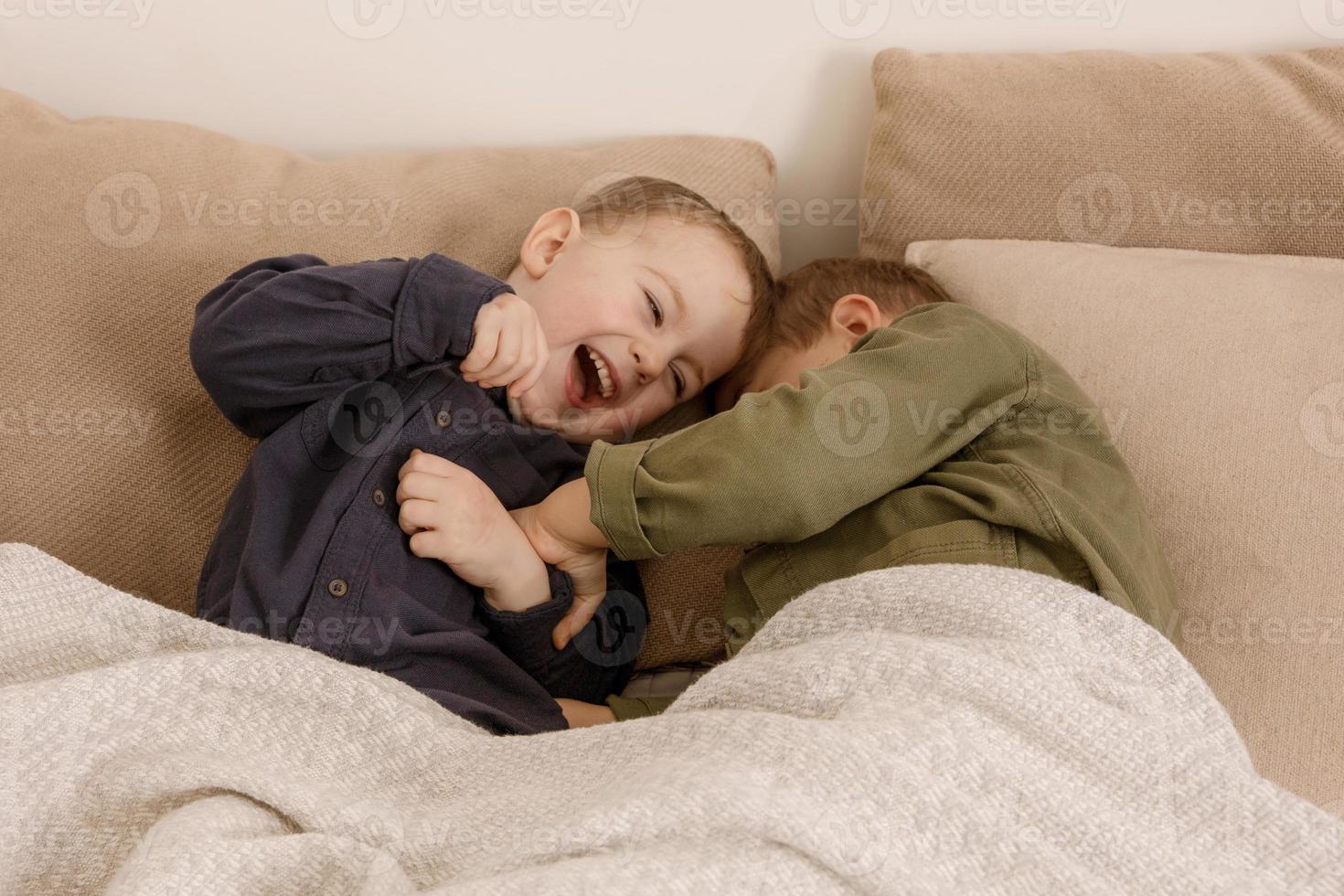 Two little and cute caucasian boys playing together on the bed at home. Interior and clothes in natural earth colors. Cozy environment. Children having fun, two brothers kittle each other. photo