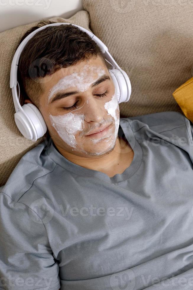 Young man with casual clothes and beauty mask on the face lying on bed at home and resting. Cosmetic for men, skin care. Man listening music or audio book. Relaxing. Time for yourself, mental health. photo