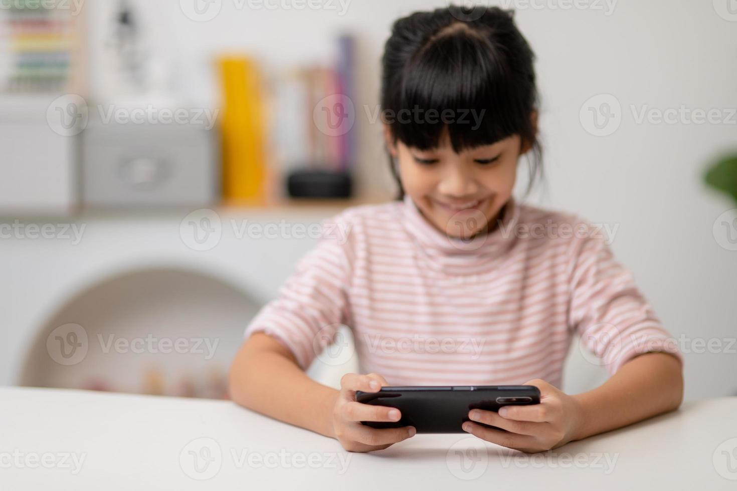 Cute little girl uses smartphone while sitting at the sofa in the living room. Child surfing the internet on mobile phone, browses through internet and watches cartoons online at home photo