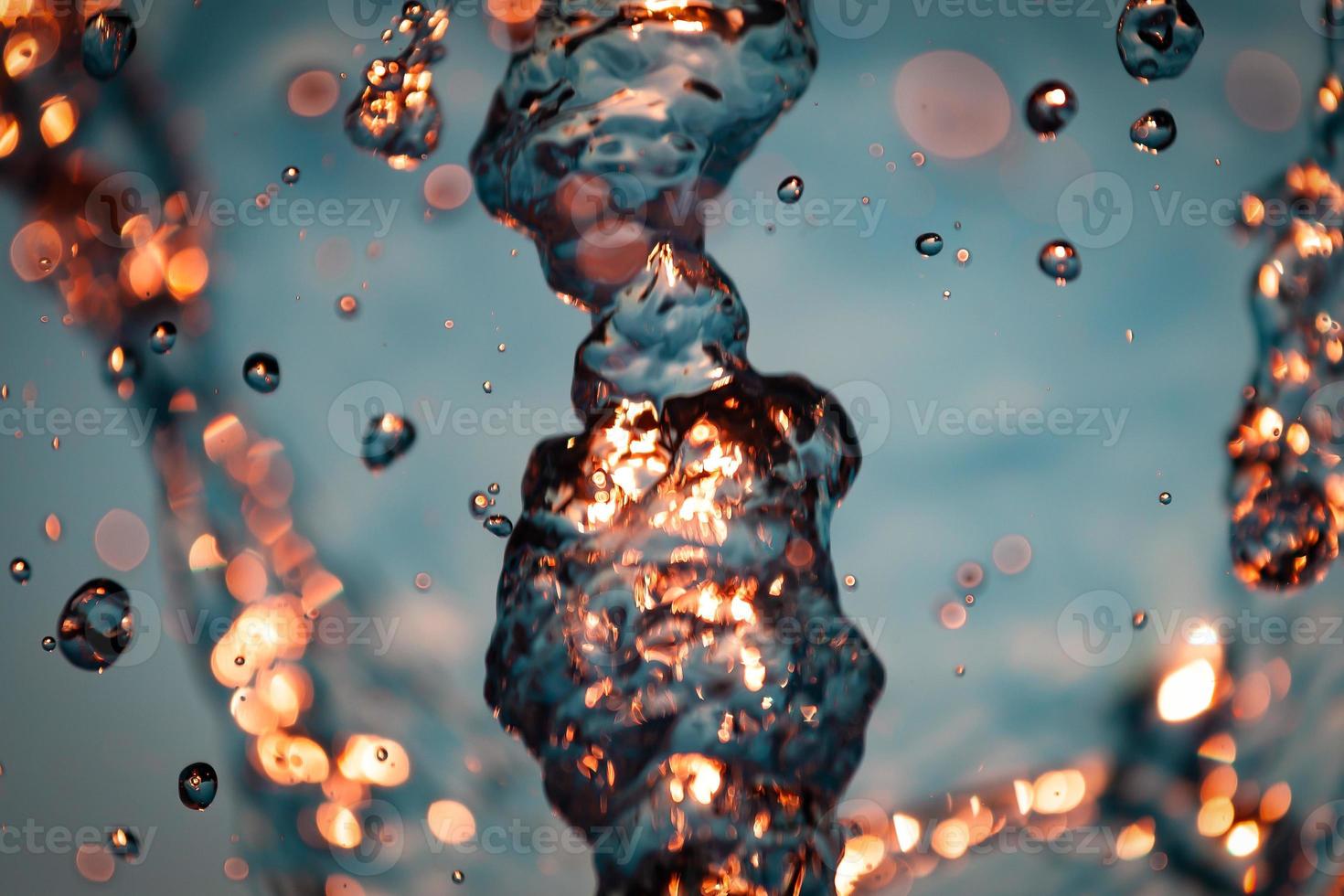 stream of water with droplets and bubbles under the orange and yellow rays of the sun. Texture of water waves of a large fountain photo