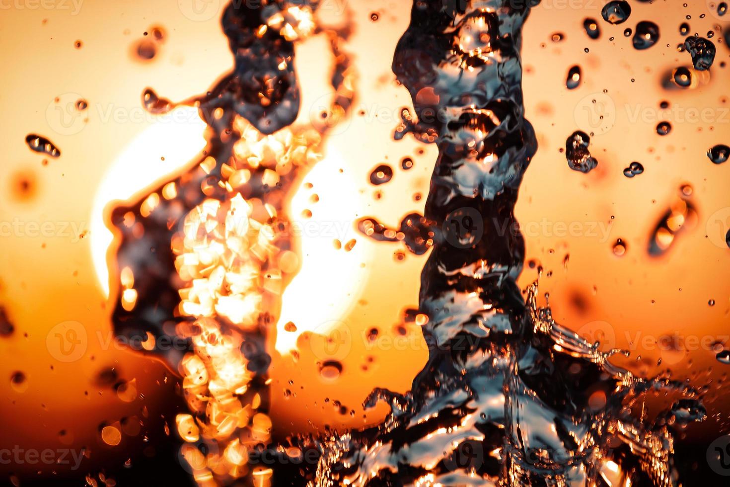 stream of water with droplets and bubbles under the orange and yellow rays of the sun. Texture of water waves of a large fountain photo