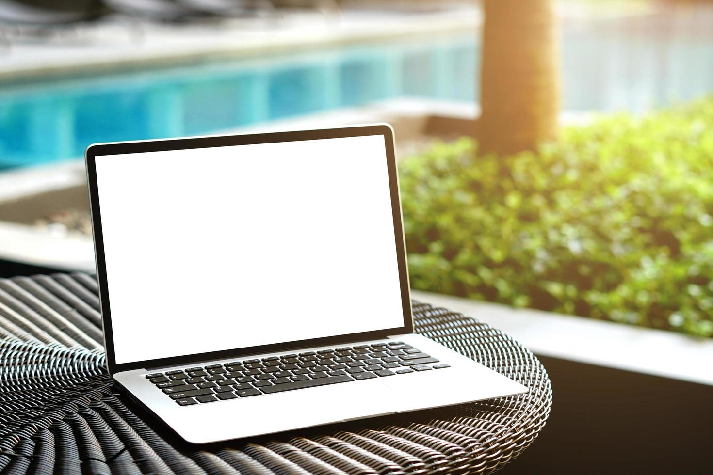 Laptop with blank screen for creative design on the table nearby pool with sun ray effect. Computer notebook with monitor clipping path for present landing page. Laptop computer mock up template photo