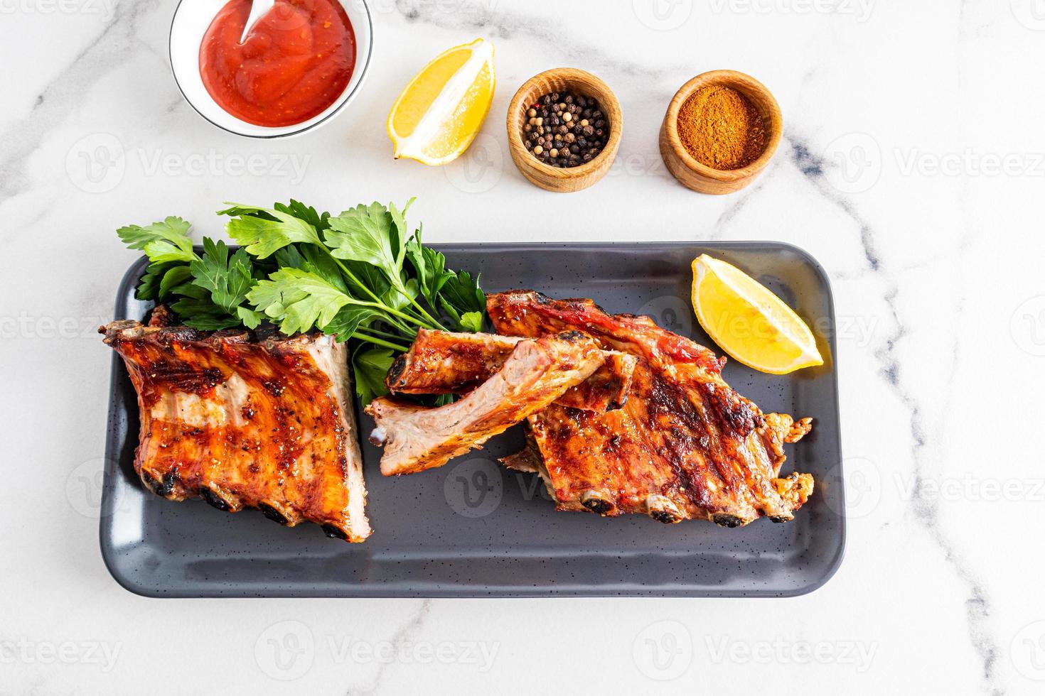 Grilled pork ribs are served on a rectangular dark ceramic plate with fresh vegetables and a bowl of sauce. white background. recipe menu. photo