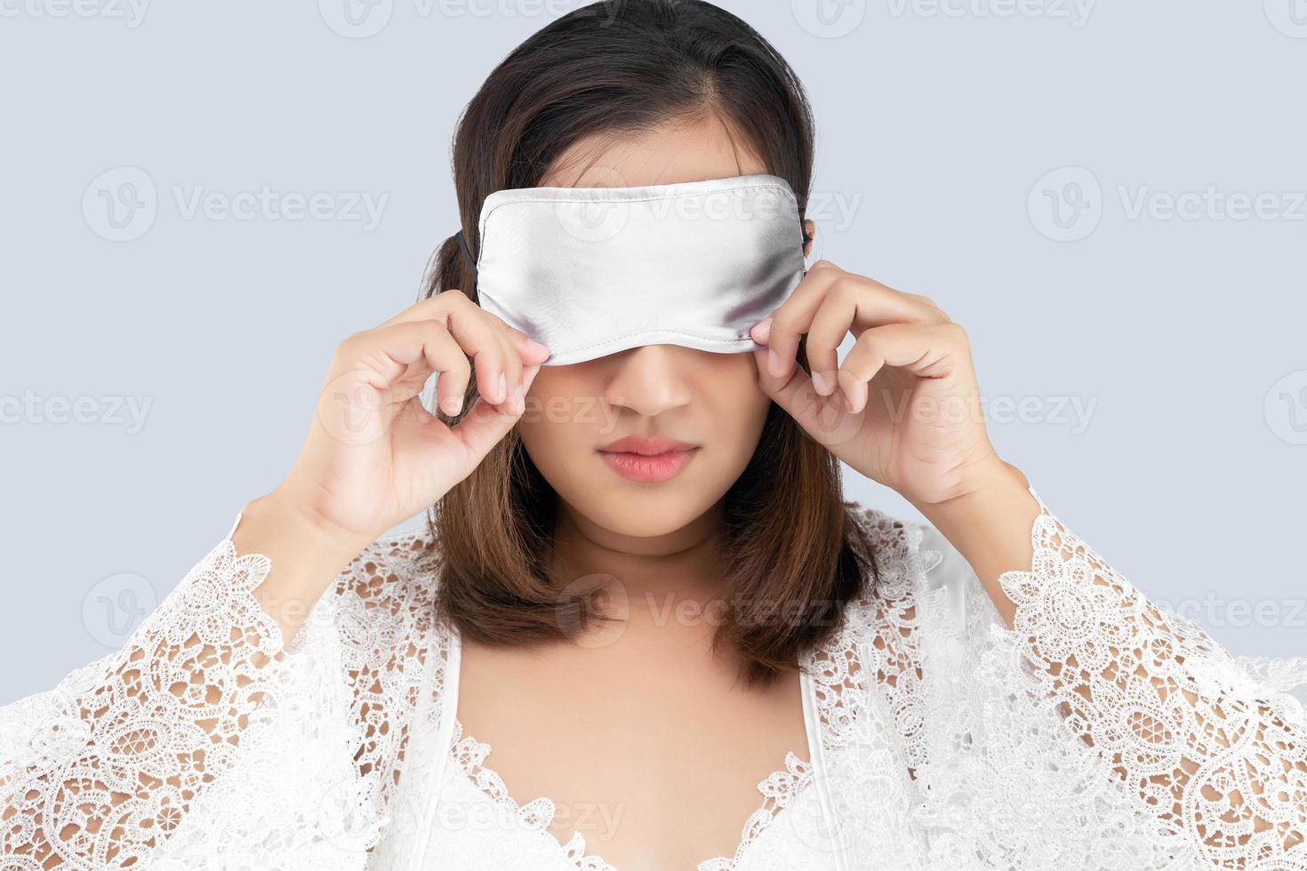 A woman in a lace nightgown and white silk robe wearing a sleep mask standing on a gray background. photo