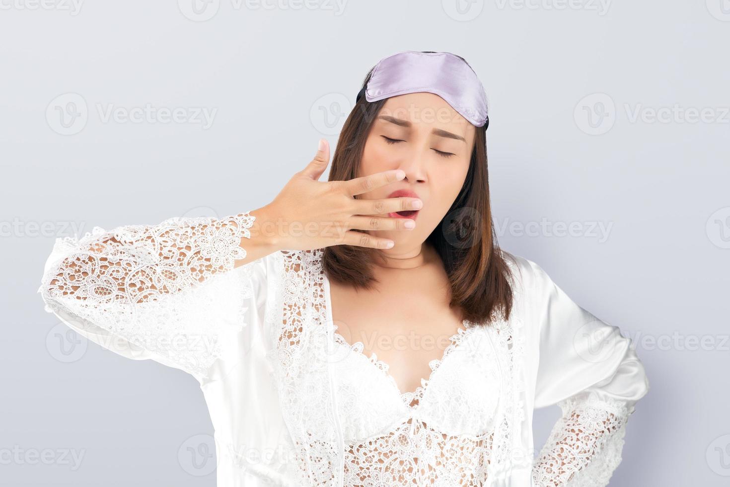 Asian woman wearing a lace white nightgown and satin robe yawning showing a sleepy gesture covering mouth with his hand. photo
