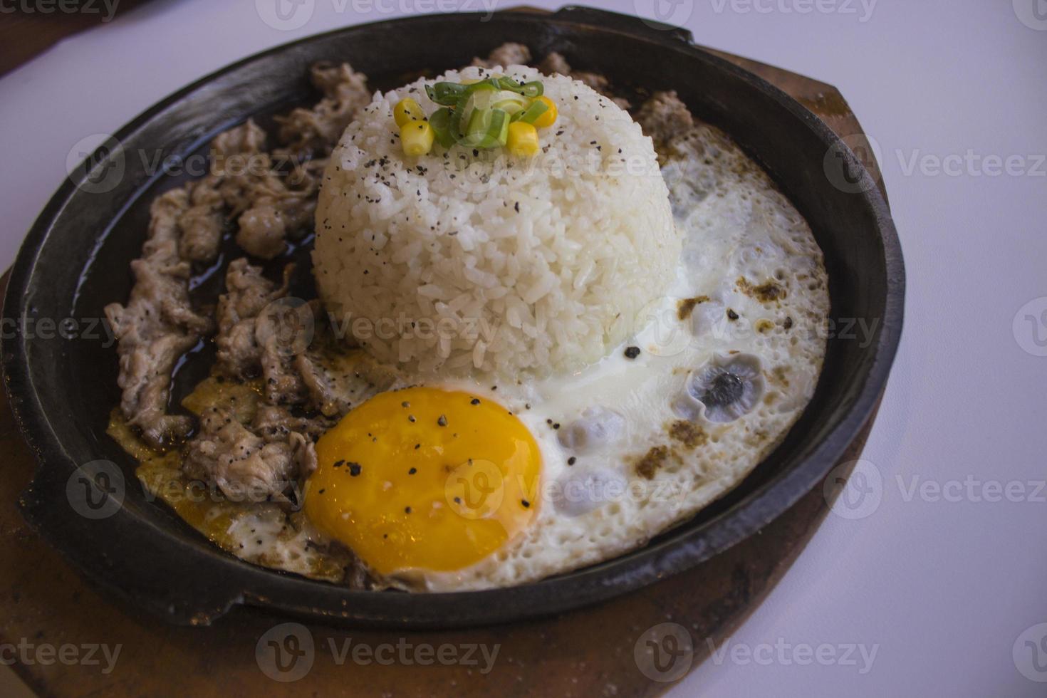 Desayuno con arroz, huevo frito y carne o ternera con guarnición de maíz y puerro en un plato caliente aislado de fondo blanco foto