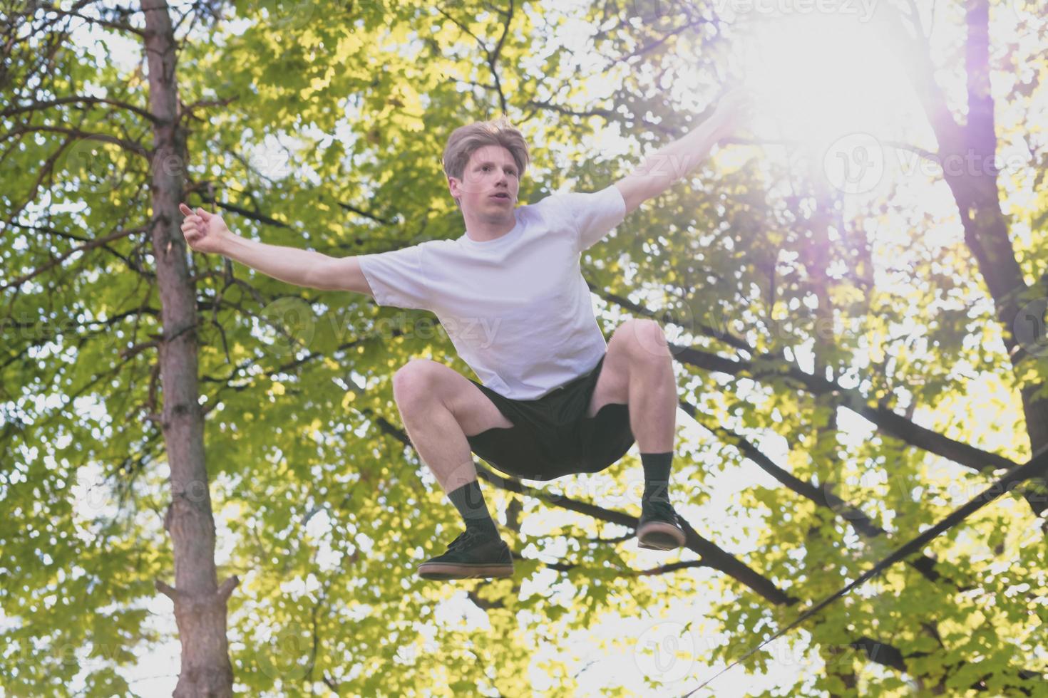 Young man balancing and jumping on slackline. Man walking, jumping and balancing on rope in park. photo