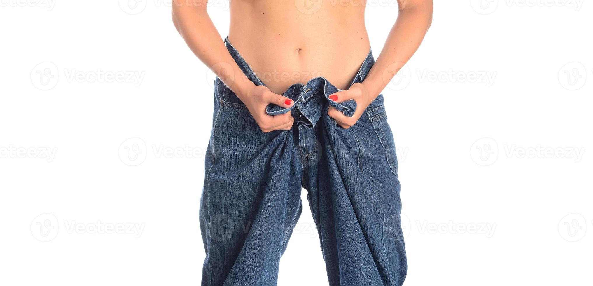 mid section of a woman with belly fat holding her blue jeans. Girl with big jeans isolated on white background. Woman shows her weight loss. Healthy lifestyle concept photo