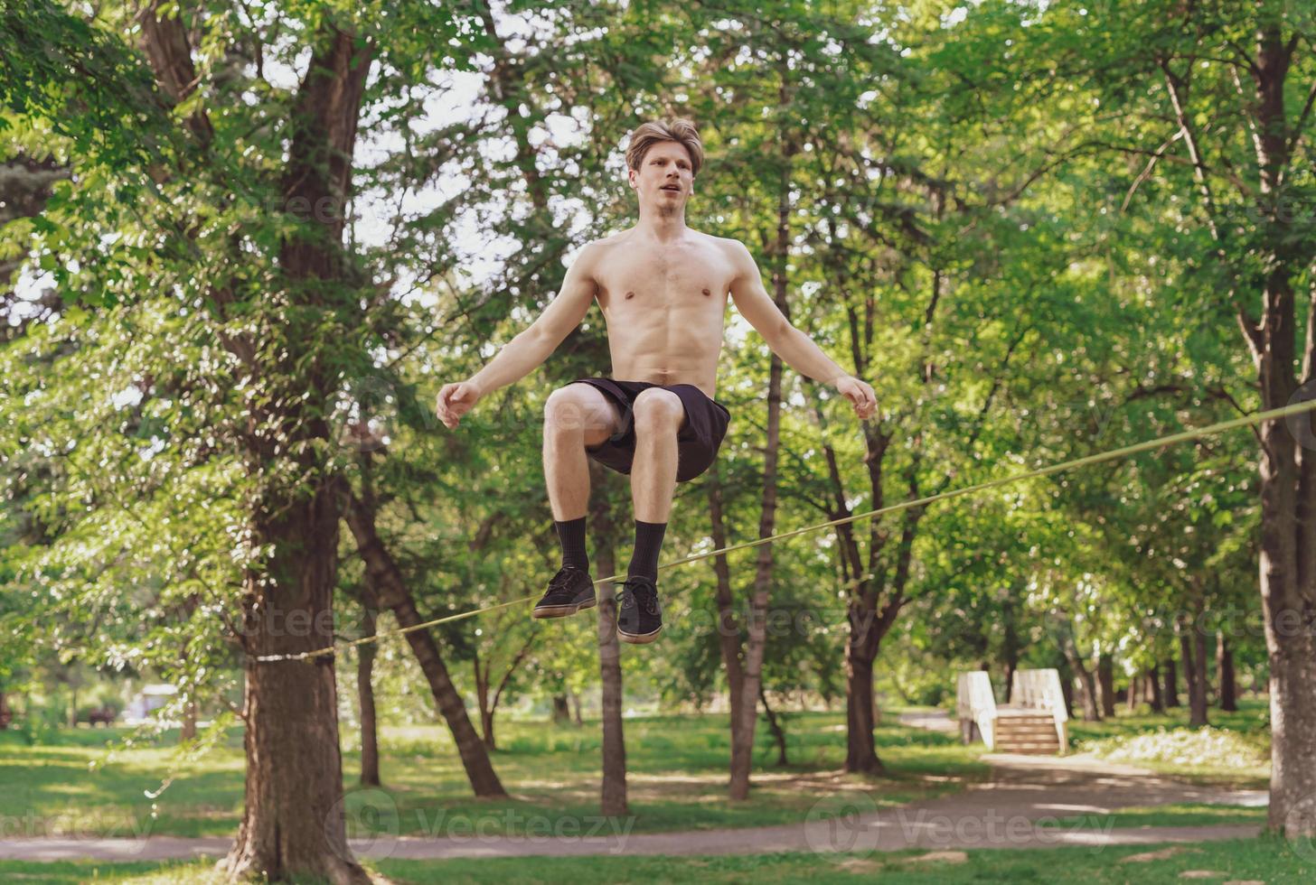 Young man balancing and jumping on slackline. Man walking, jumping and balancing on rope in park. photo