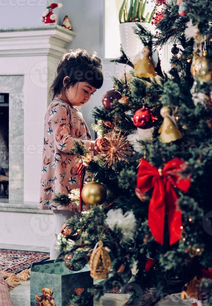Asian little girl decorate the Christmas tree indoors. The morning before Xmas. Portrait loving girl close up. photo