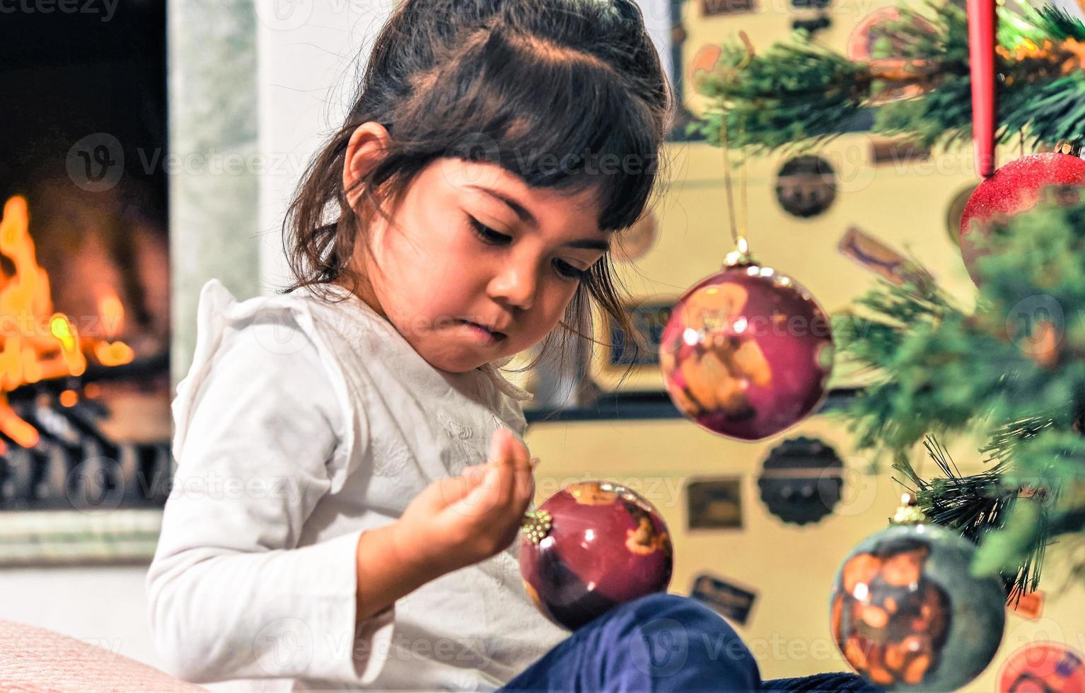 Asian little girl decorate the Christmas tree indoors. The morning before Xmas. Portrait loving girl close up. photo