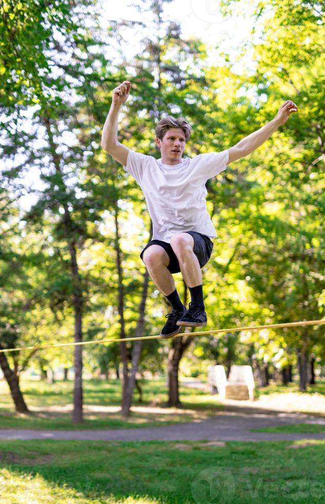 Young man balancing and jumping on slackline. Man walking, jumping and balancing on rope in park. photo