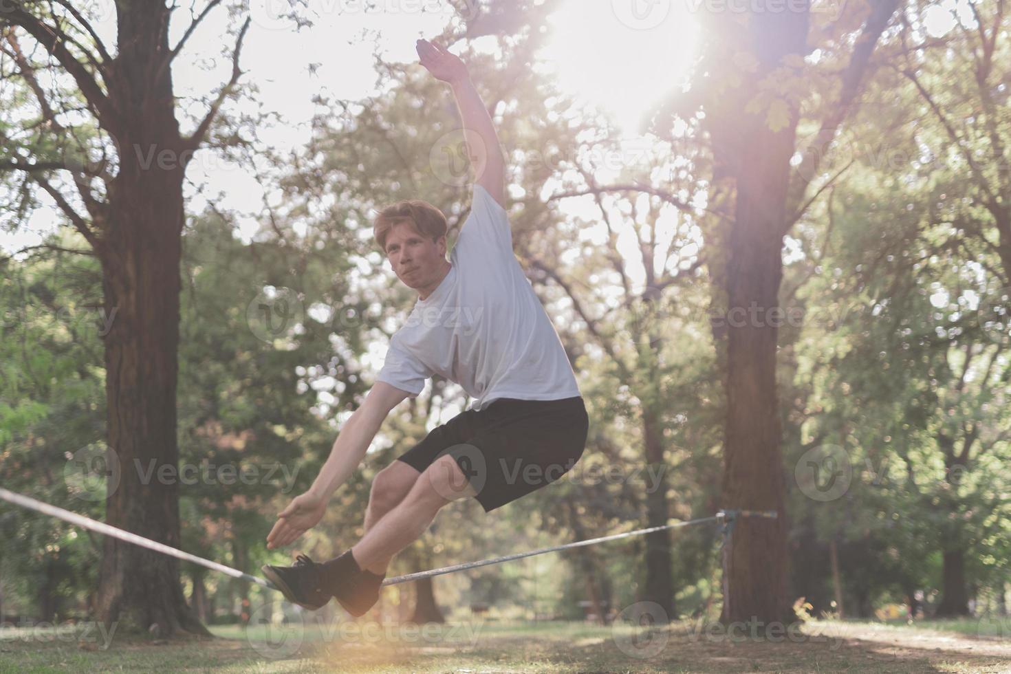 Young man balancing and jumping on slackline. Man walking, jumping and balancing on rope in park. photo