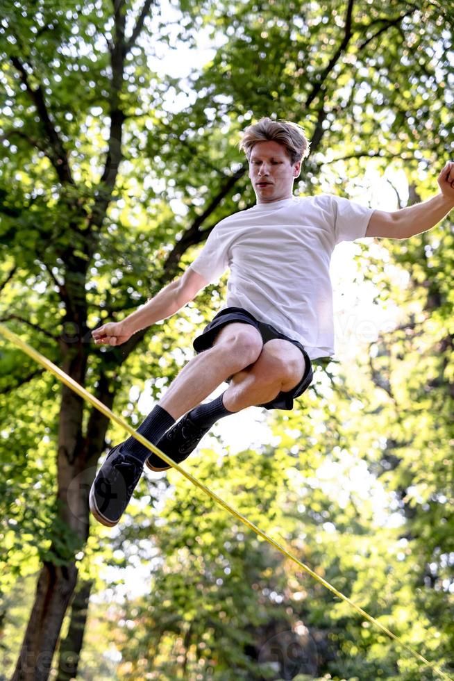 Young man balancing and jumping on slackline. Man walking, jumping and balancing on rope in park. photo