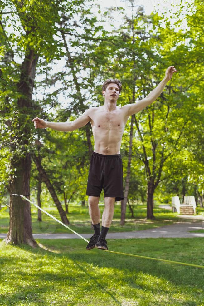 Young man balancing and jumping on slackline. Man walking, jumping and balancing on rope in park. photo