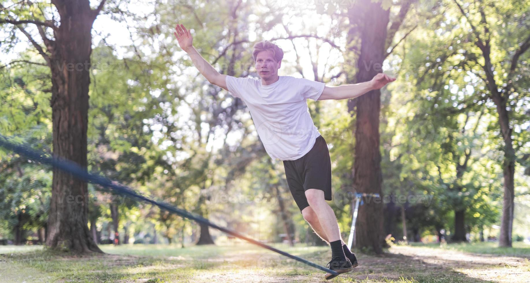 Young man balancing and jumping on slackline. Man walking, jumping and balancing on rope in park. photo