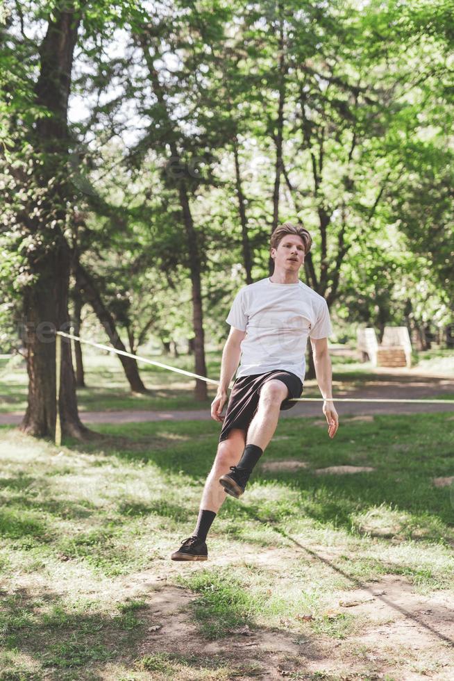 Young man balancing and jumping on slackline. Man walking, jumping and balancing on rope in park. photo