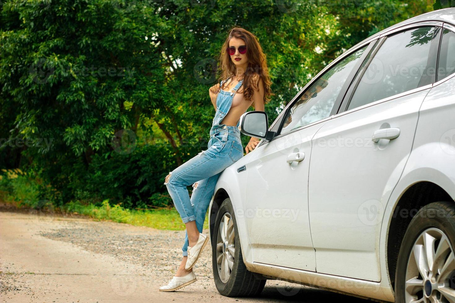 Sexy young brunette in jeans overall stans across a white car photo