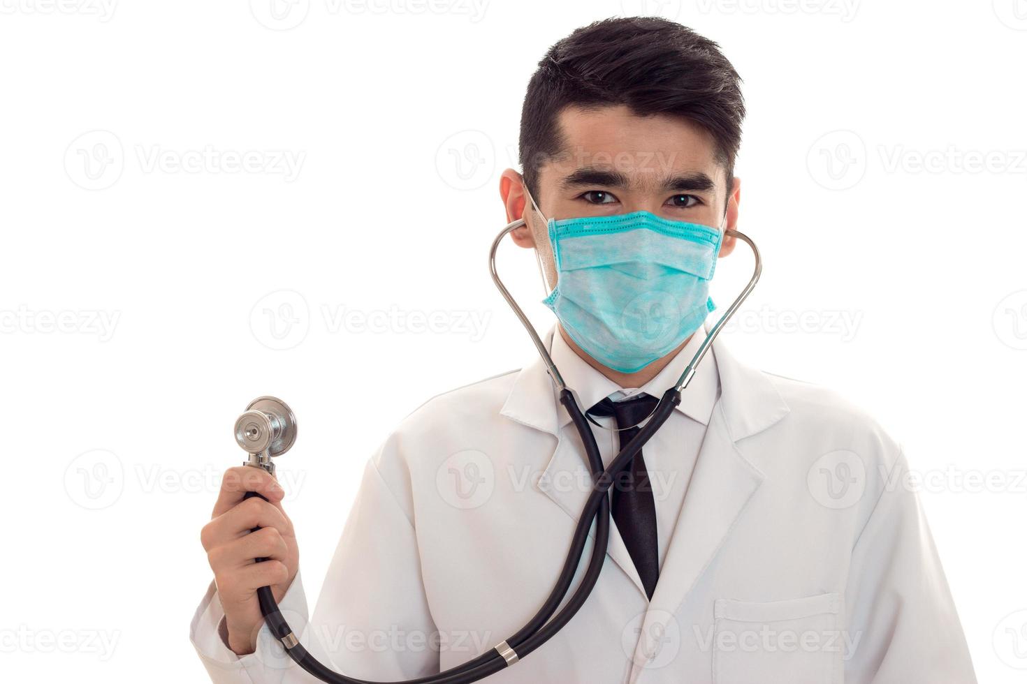 retrato de un joven moreno doctor que me enmascara con estetoscopio mirando la cámara aislada de fondo blanco foto