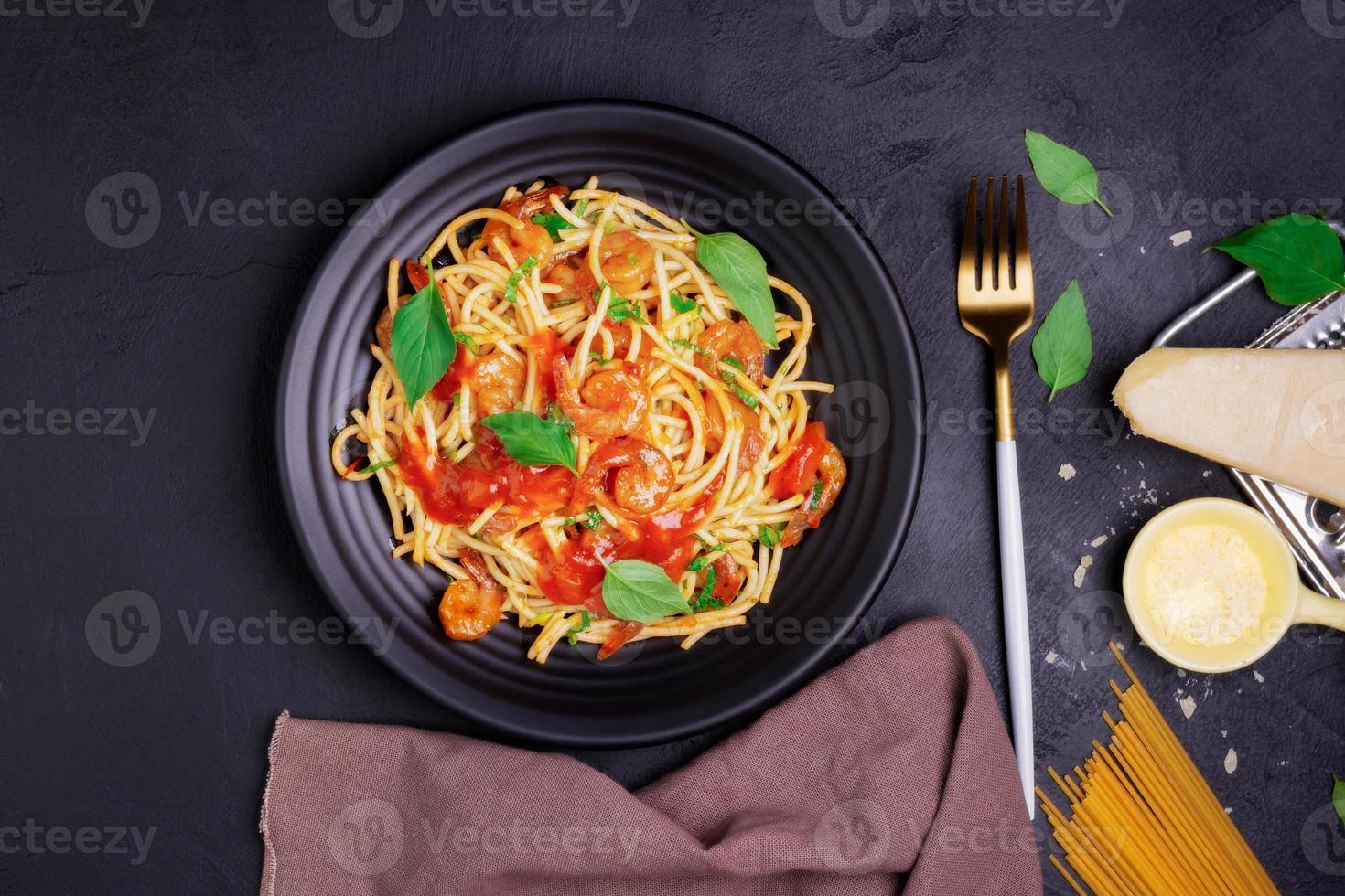 Delicious spaghetti pasta with prawns and cheese served on a black plate. With vegetables, Italian tomato sauce, and spices arranged on a wooden table, black background, top view photo
