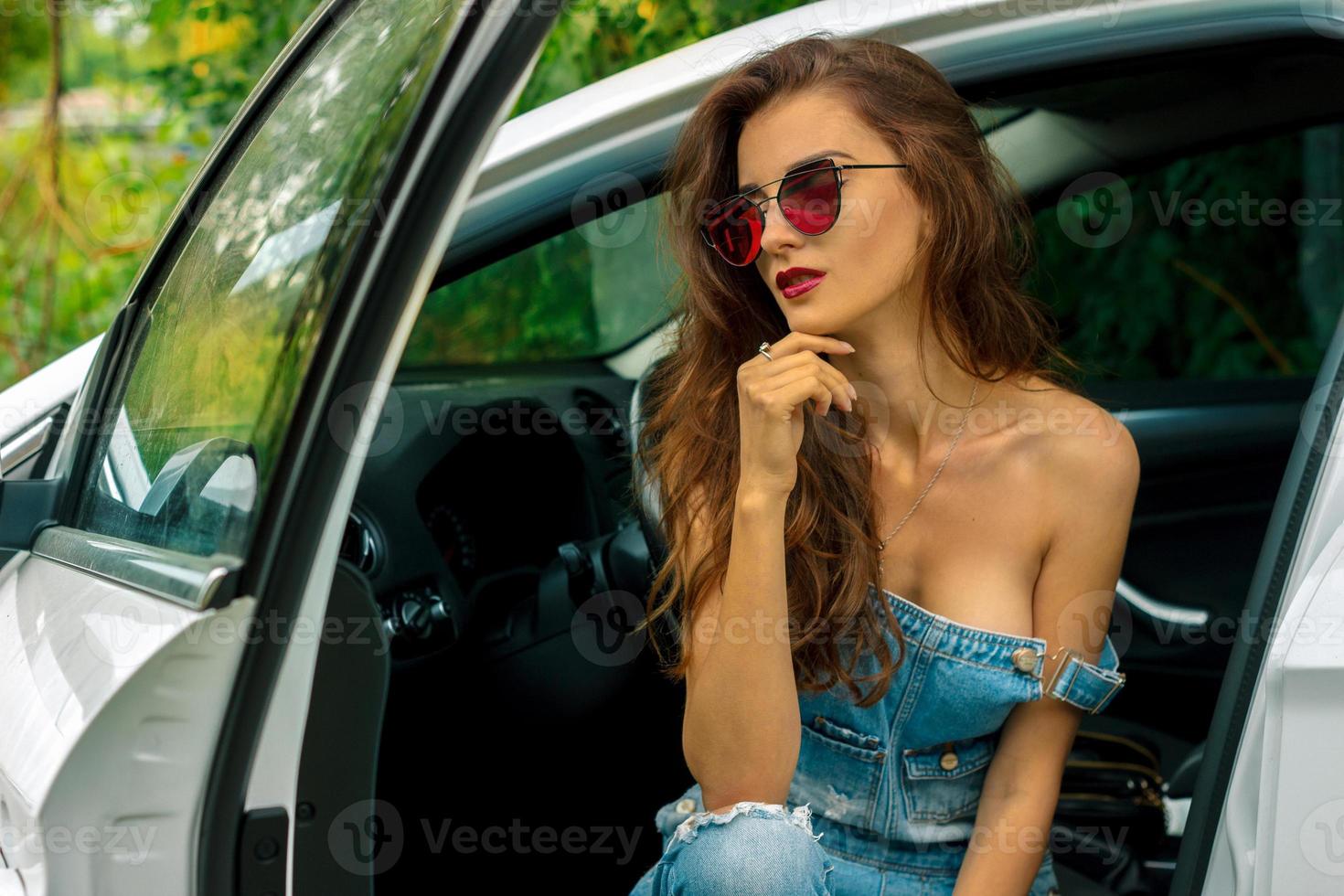 gorgeous young brunette girl with sexy body in jeans overall sits in a car photo