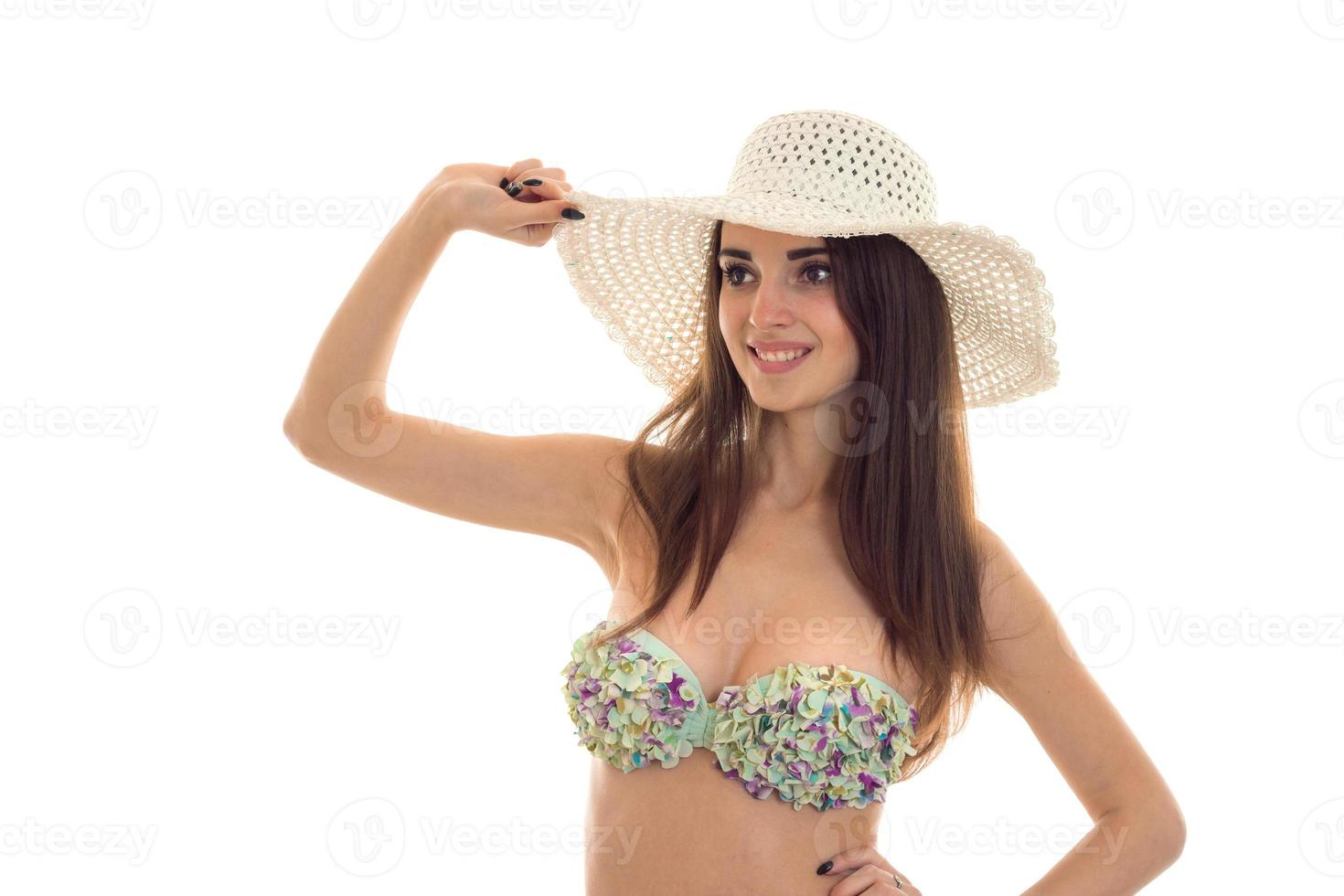 young cheerful brunette woman with big natural breasts in swimsuit with floral pattern and straw hat smiling and looking away isolated on white background photo