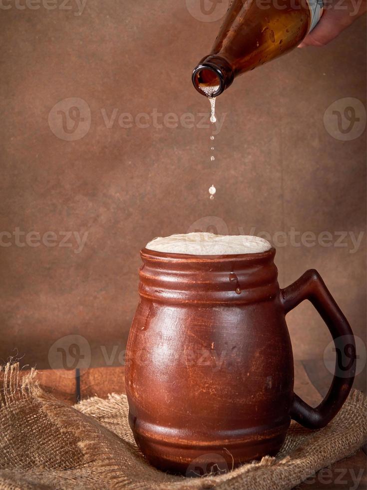 Beer clay brown mug with beer on a wooden table on a dark background. A stream of light beer pours into a glass photo