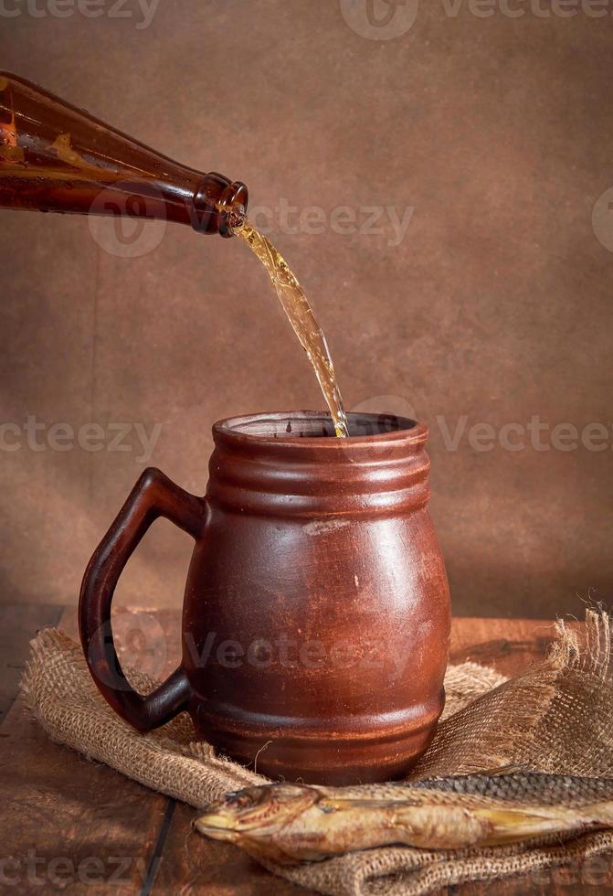 Beer clay brown mug with beer on a wooden table on a dark background. A stream of light beer pours into a glass photo