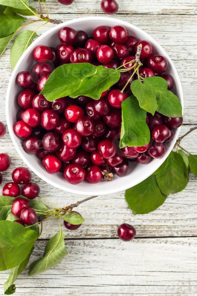 tazón blanco de cerezas dulces frescas con hojas en gotas de agua sobre fondo de madera foto