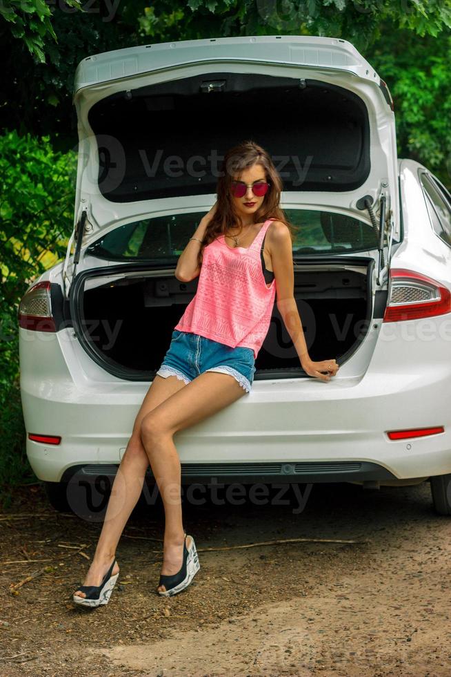 chica de moda en pantalones cortos y una camisa brillante se sienta en el maletero de un coche foto