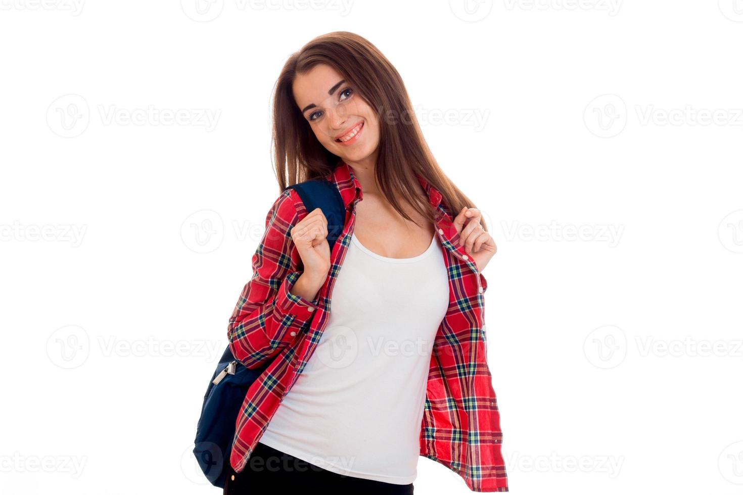 alegre joven estudiante morena con mochila azul posando y mirando a la cámara y sonriendo aislada en el fondo blanco foto