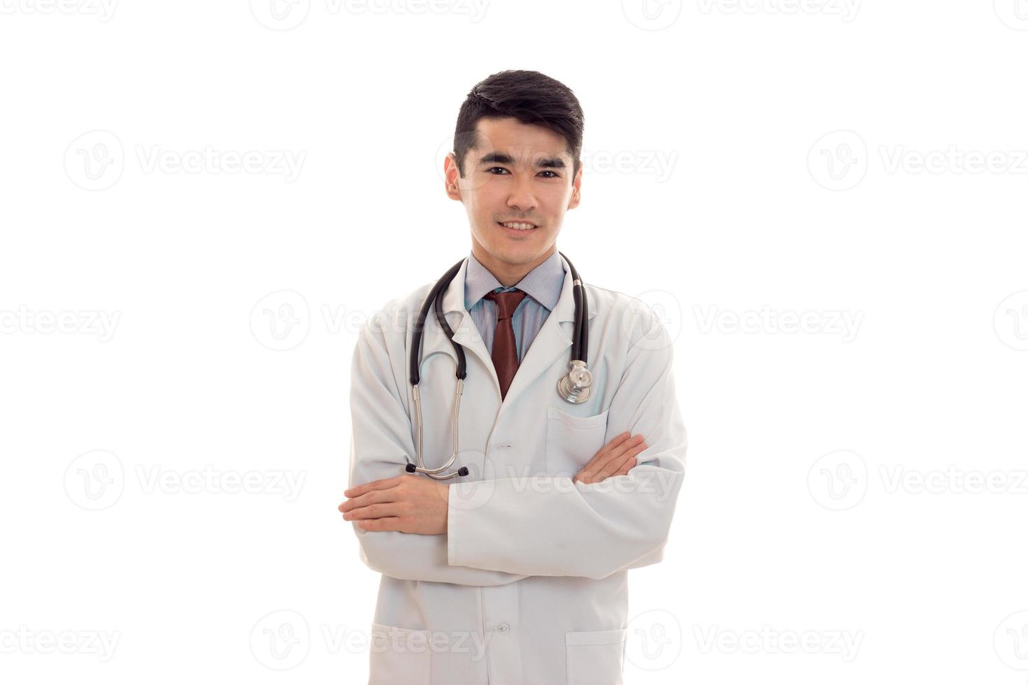 pretty young brunette man doctor in white uniform with stethoscope looking and smiling on camera isolated in studio photo