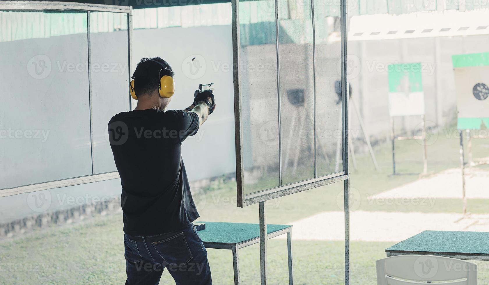 tirador asiático con auriculares con cancelación de ruido y ropa negra practicando disparar armas cortas en el campo de tiro. Tiro deportivo para meditación y defensa personal, actividades recreativas. foto