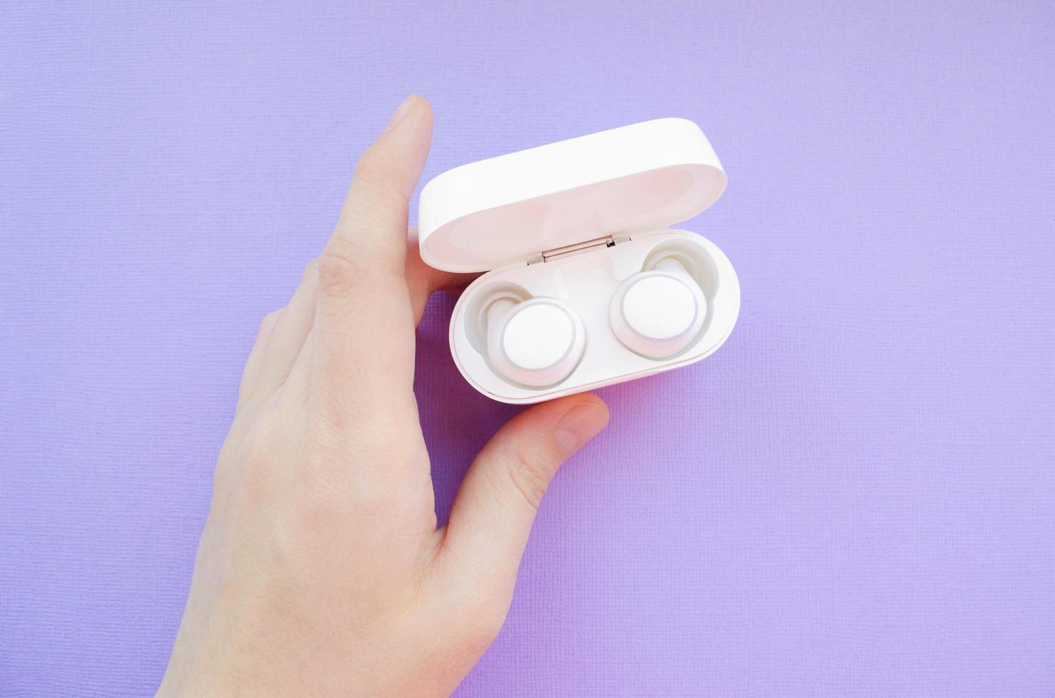 Hand holds wireless headphones case. White true wireless earbuds. Wireless earbuds on purple background. photo