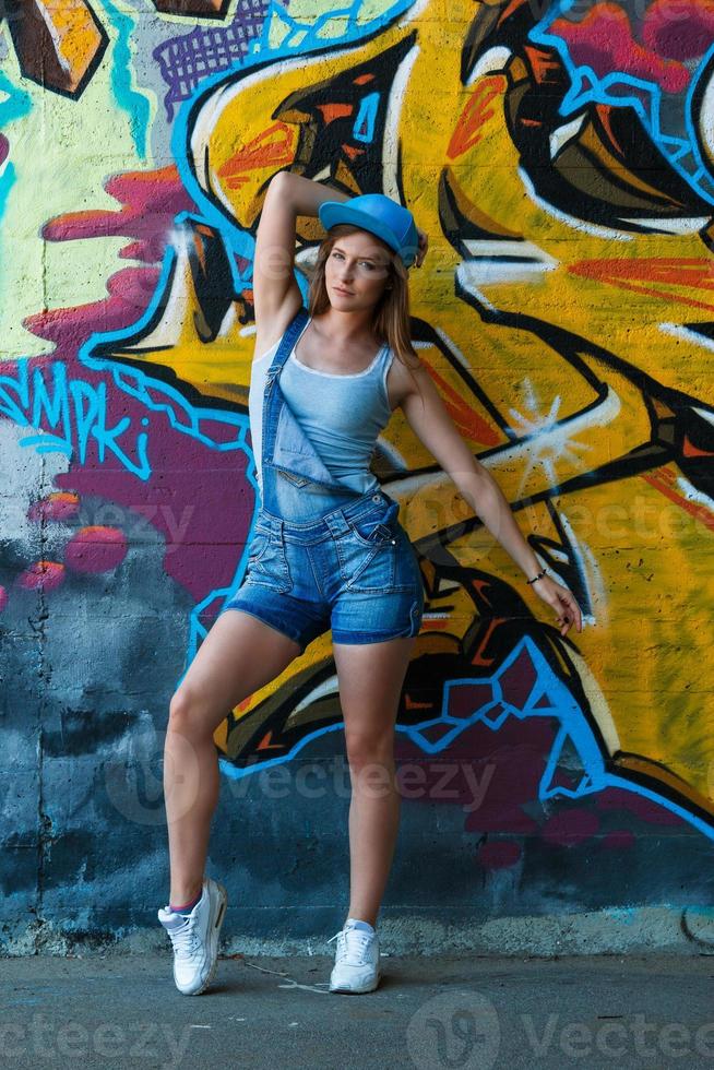 Girl in denim overalls posing against wall with graffiti photo