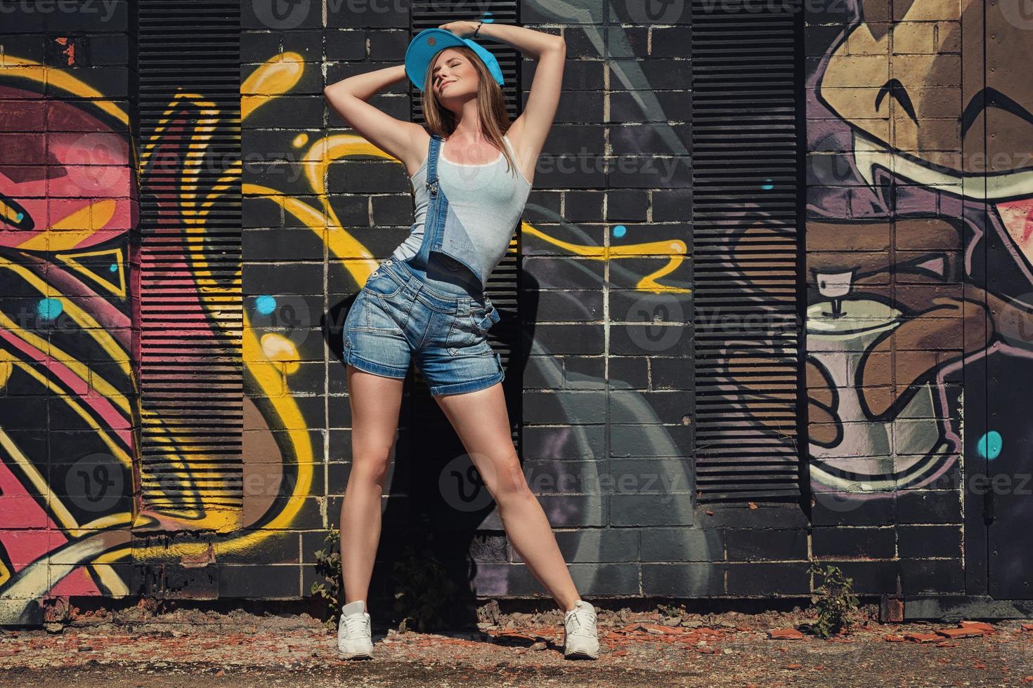 Girl in denim overalls posing against wall with graffiti photo