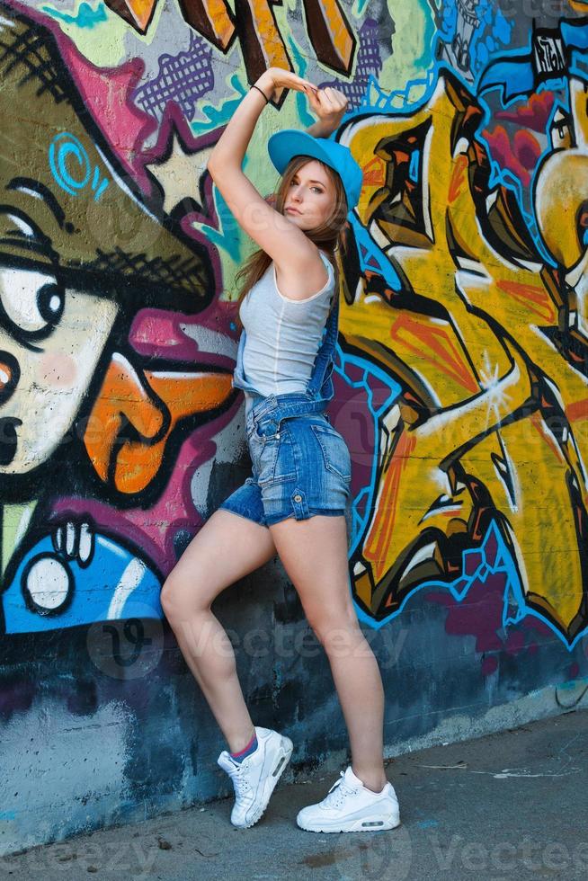 Girl in denim overalls posing against wall with graffiti photo