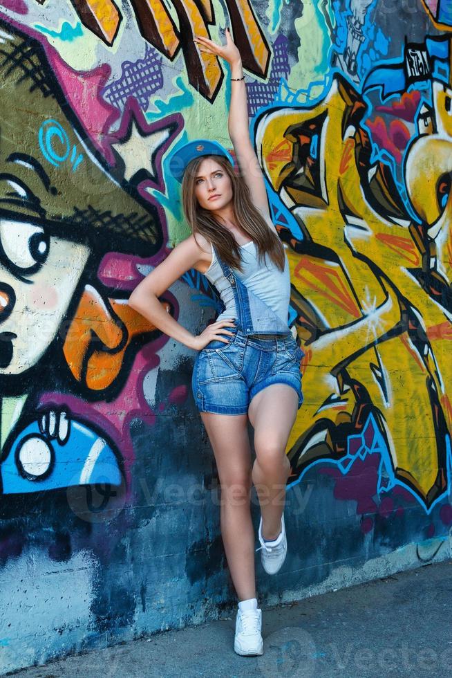 Girl in denim overalls posing against wall with graffiti photo