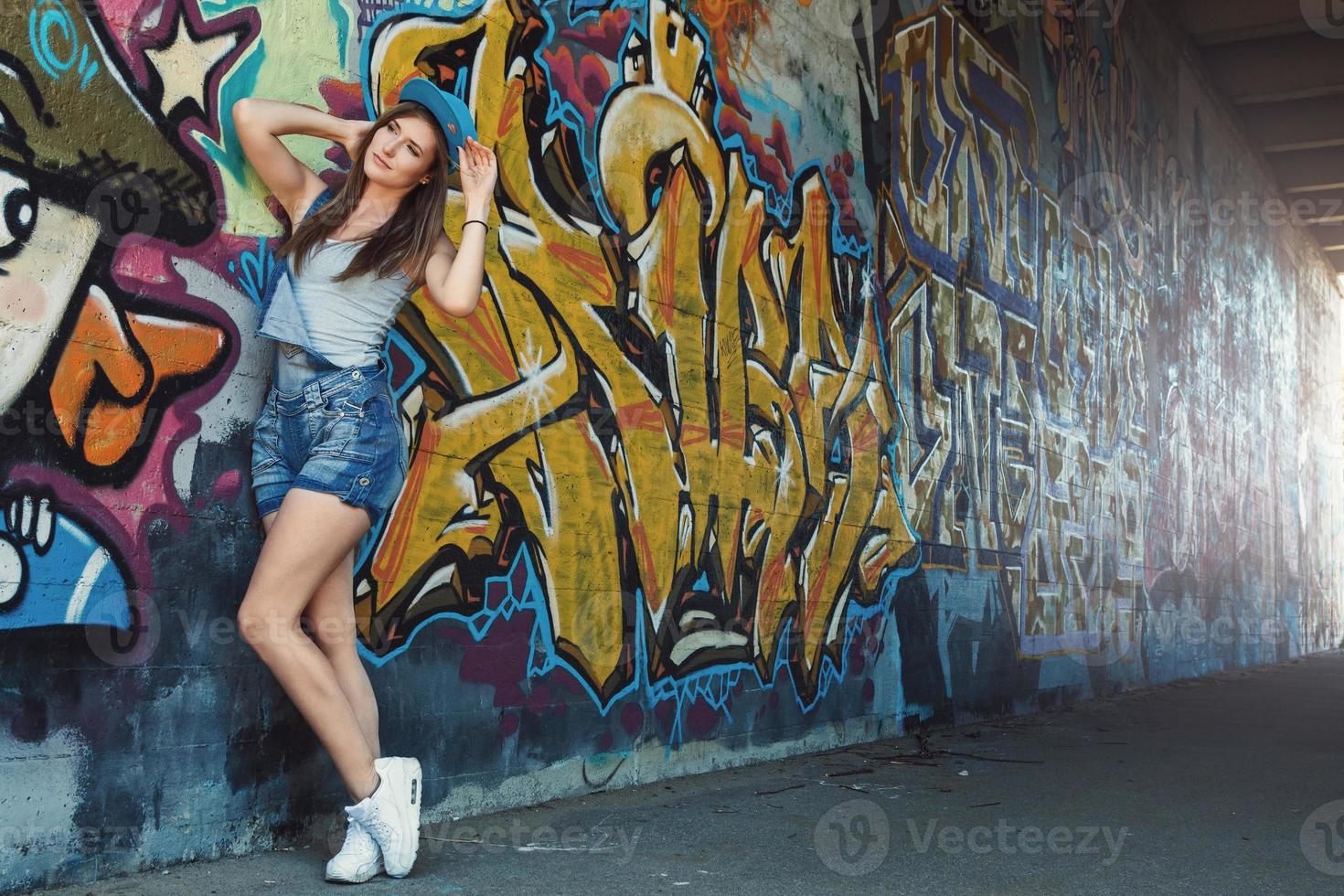 Girl in denim overalls posing against wall with graffiti photo