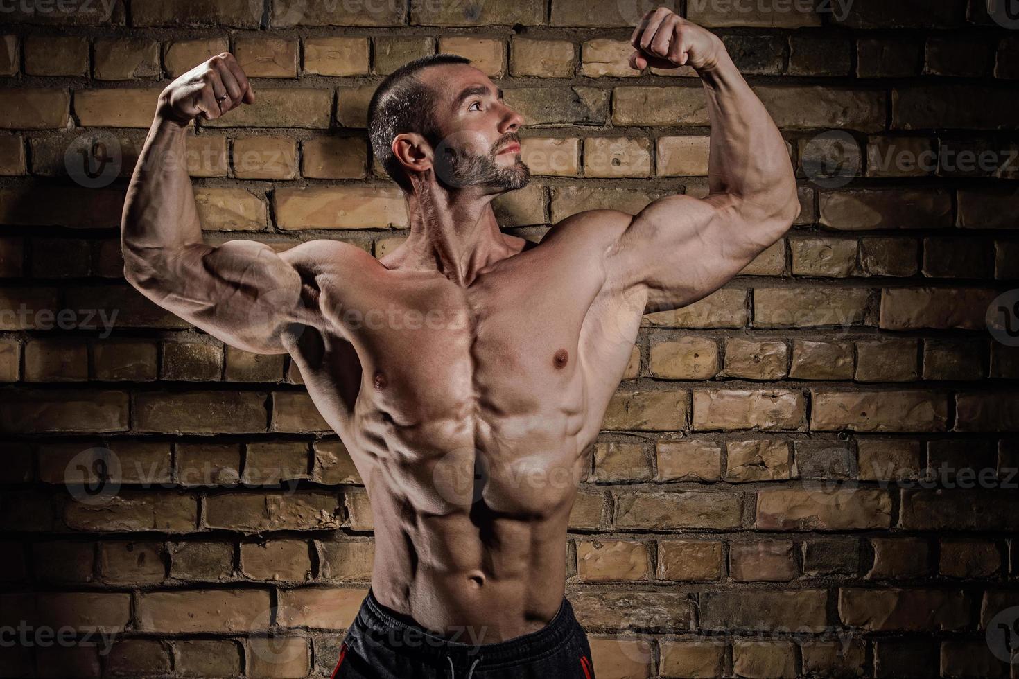 Bodybuilder showing his muscles against brick wall photo