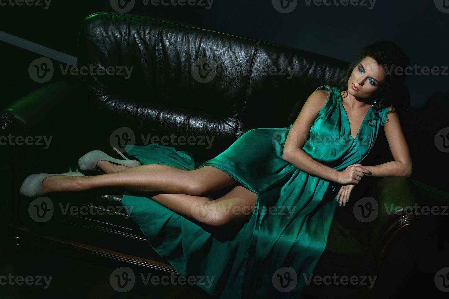 Woman wearing beautiful green silk dress is posing on leather couch photo