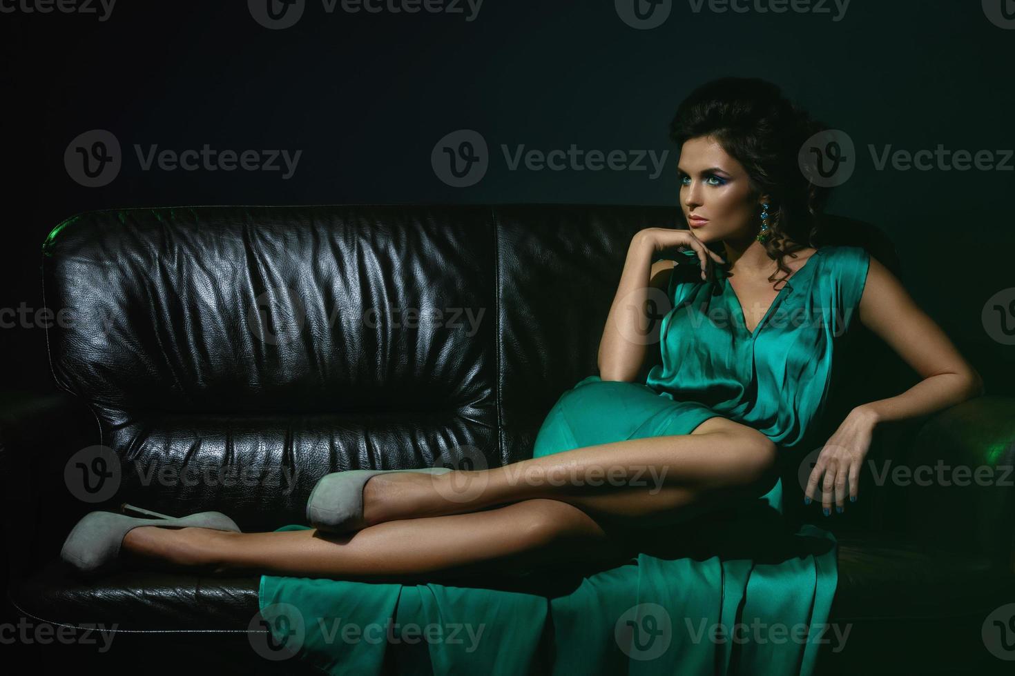Woman wearing beautiful green silk dress is posing on leather couch photo
