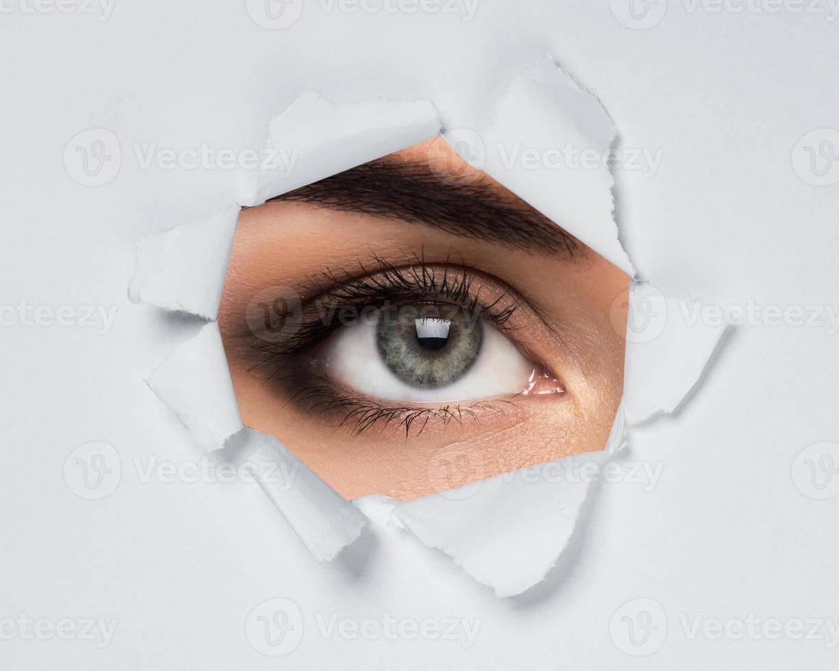 Young female eye looking through torn paper photo
