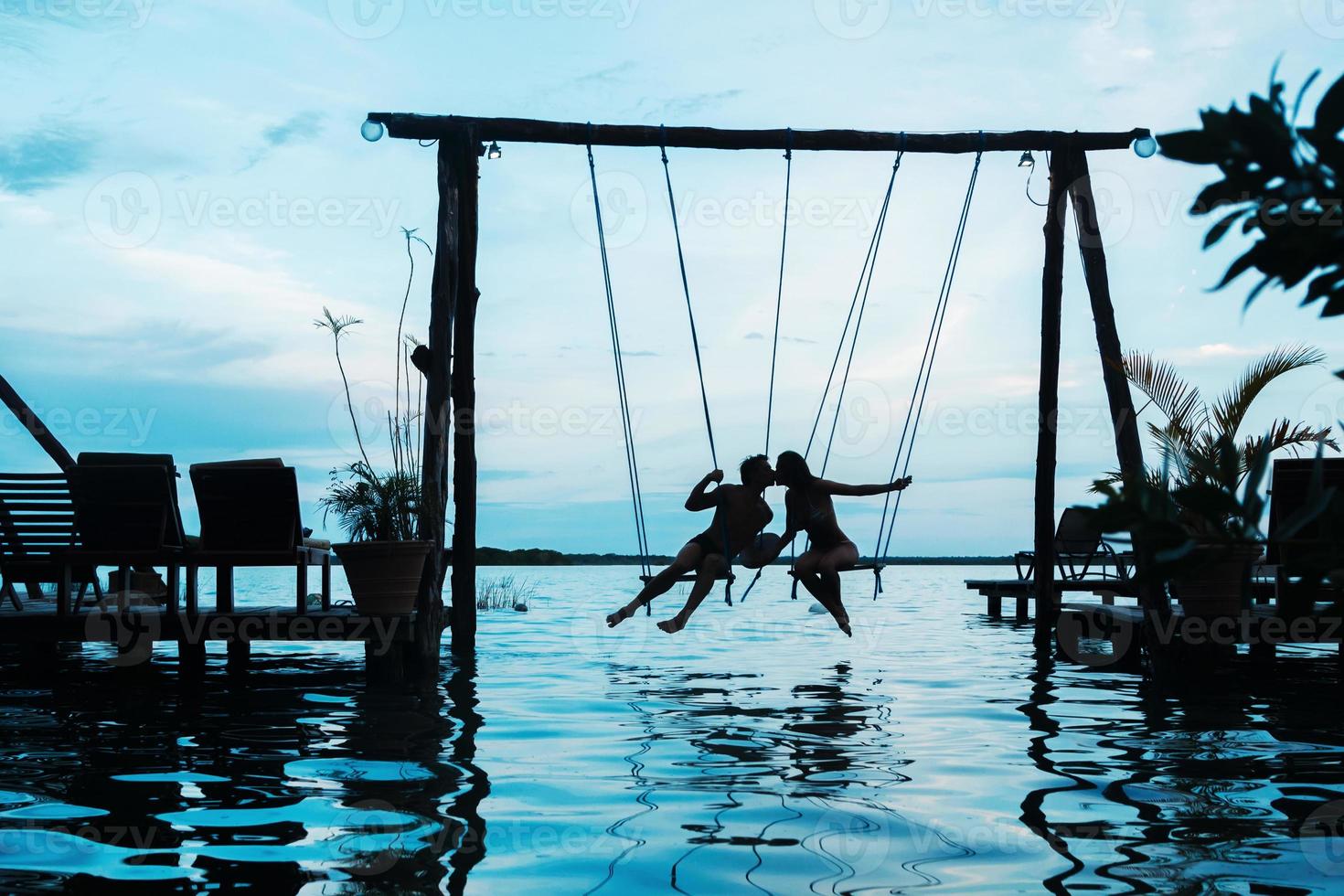 Silhouette of couple on swings which is above the water photo