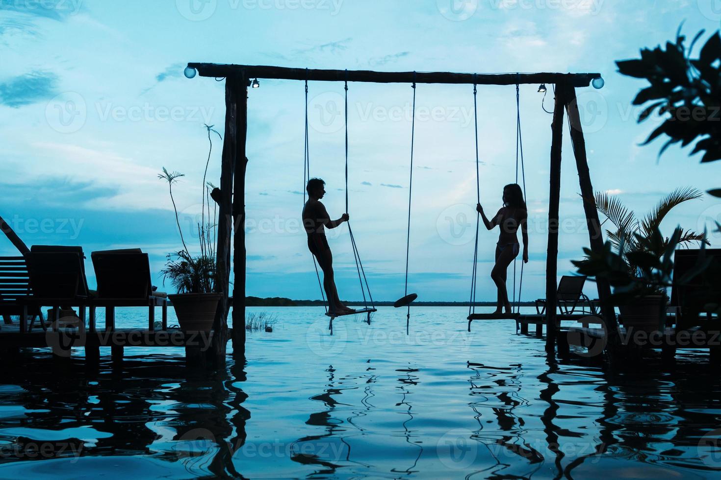 Silhouette of couple on swings which is above the water photo