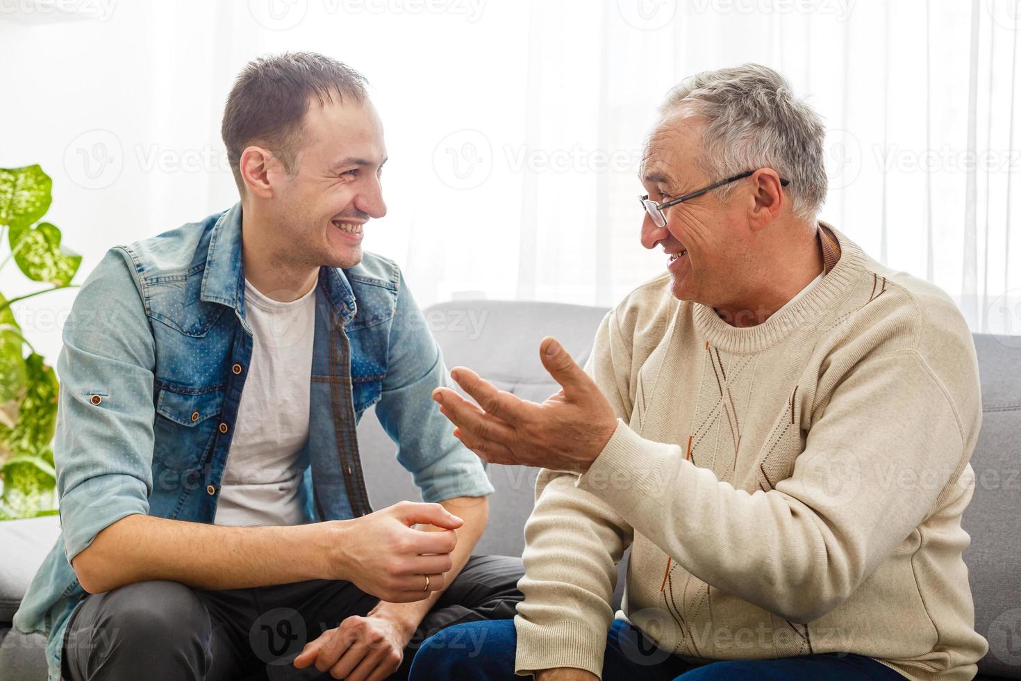 Smiling grown son sit on couch relax with senior dad talk sharing thoughts looking in eyes, happy millennial man rest on sofa speak with elderly father enjoy leisure family weekend at home. photo