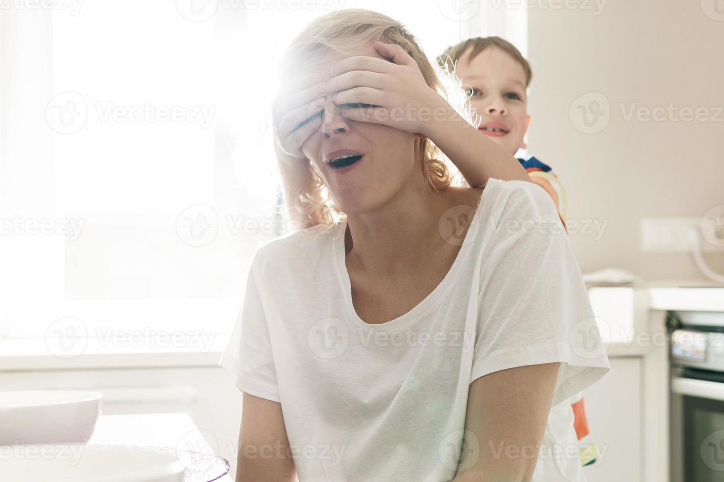 chico lindo está cubriendo los ojos de su madre en la cocina foto