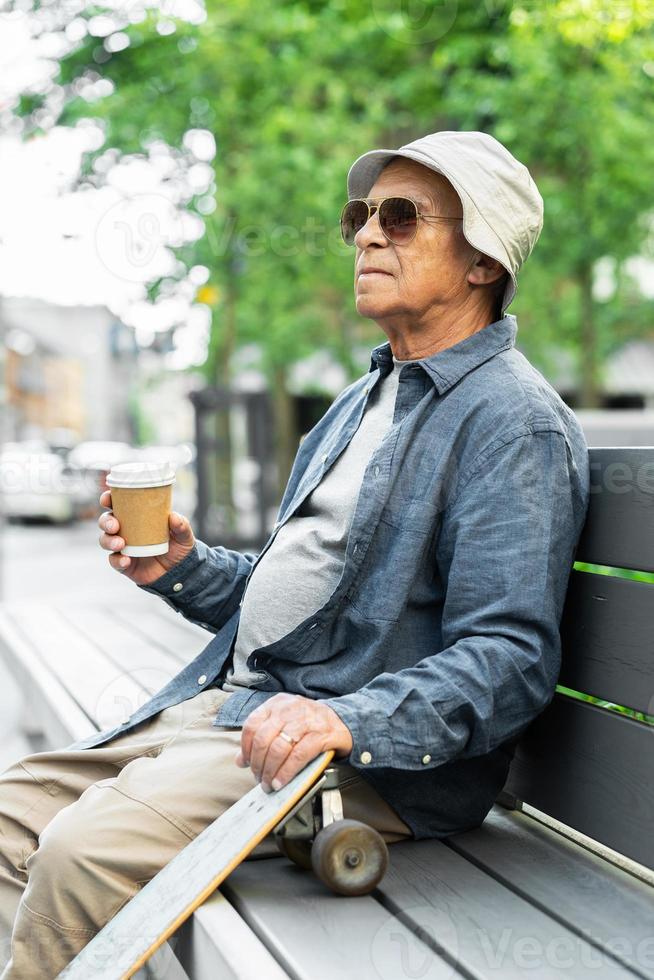 Elderly man with a longboard sitting on the bench and drinking coffee in a city park photo