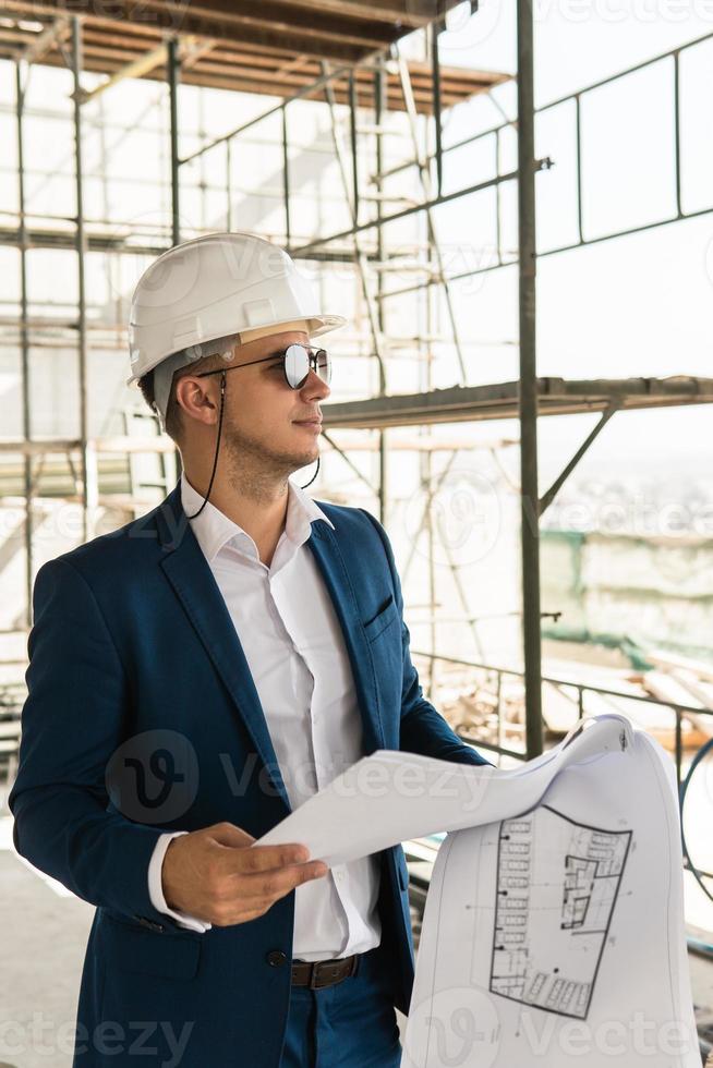 Arquitecto hombre vestido con traje formal y sombrero duro durante el control de la construcción de edificios sosteniendo planos en un sitio de construcción foto