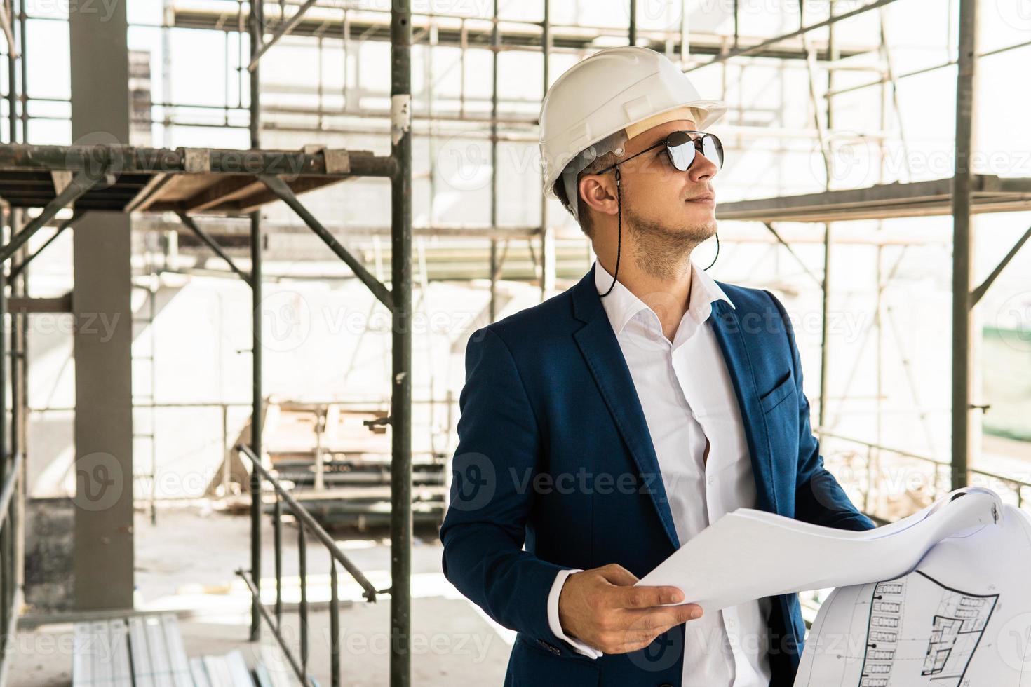 Man architect wearing formal suit and hard hat during building construction control holding a blueprints on a construction site photo